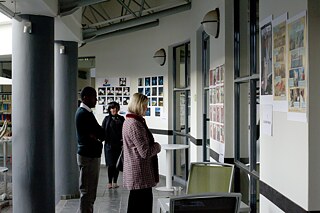 Das Bild zeigt drei Personen, die im seitlichen Umlauf des Atriums stehen und verschiedene Kunstwerke oder Poster an den Wänden betrachten. Die Wände sind mit mehreren Bildern in einem Rastermuster geschmückt. Es gibt auch einige Tische und Stühle, die dort vereinzelt stehen.